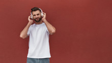 Favourite Playlist. Portrait Of Handsome Man In Headphones Listening To The Music And Looking At Camera While Standing Against Red Wall Outdoors