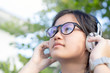Portrait of asian young girl wearing glasses,headphones listening to music at green nature outdoor background.Closeup face of teen hand holding wearing headphone and keeps the rhythm of the song.