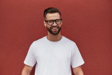 I Am Happy! Portrait Of Cheerful Attractive Bearded Man In Eyeglasses Smiling And Looking At Camera While Standing Against Red Wall Outdoors