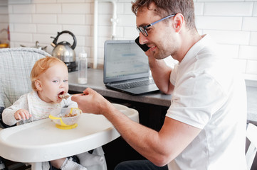 Happy young father working at home and sitting with a baby girl. Father feeding a baby during talking the mobile phone