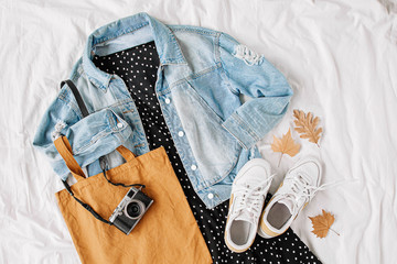 Wall Mural - Blue jean jacket and black dress, sneakers with tote bag and photo camera on white bed. Women's stylish autumn or spring outfit. Trendy clothes. Flat lay, top view.