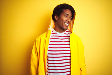 Sticker - African man with afro hair wearing rain coat with hood standing over isolated yellow background looking away to side with smile on face, natural expression. Laughing confident.