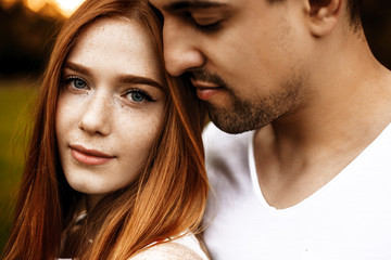 Wall Mural - Close up portrait of a beautiful caucasian young couple embracing while she is looking at camera smiling and he is smelling her hair with closed eyes outside.