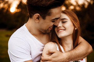 Wall Mural - Young caucasian smiling man embracing from back his girlfriend with freckles and red hair which is with closed eyes smiling against sunset.