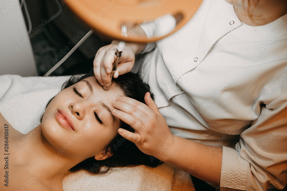 Upper view portrait of a lovely caucasian female having non invasive microdermabrasion on her face while laying in a spa bed with closed eyes. - obrazy, fototapety, plakaty 