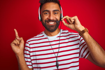 Wall Mural - Young indian call center agent man using headset over isolated red background very happy pointing with hand and finger to the side