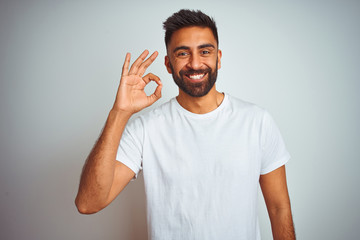 Wall Mural - Young indian man wearing t-shirt standing over isolated white background smiling positive doing ok sign with hand and fingers. Successful expression.