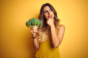 Wall Mural - Young beautiful woman eating broccoli over yellow isolated background serious face thinking about question, very confused idea