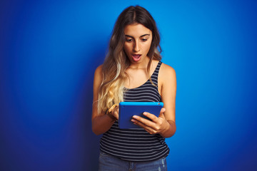 Poster - Young beautiful woman using touchpad tablet over blue isolated background scared in shock with a surprise face, afraid and excited with fear expression