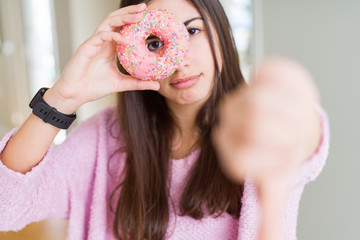 Sticker - Beautiful young woman eating pink chocolate chips donut with angry face, negative sign showing dislike with thumbs down, rejection concept