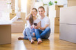 © Krakenimages.com - Beautiful couple sitting on the floor using smartphone at new home around cardboard boxes with happy face smiling doing ok sign with hand on eye looking through fingers