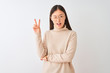 © Krakenimages.com - Young chinese woman wearing turtleneck sweater and glasses over isolated white background smiling with happy face winking at the camera doing victory sign. Number two.