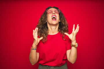 Sticker - Middle age senior woman wearing cute heart shaped glasses over red isolated background crazy and mad shouting and yelling with aggressive expression and arms raised. Frustration concept.