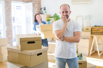 Sticker - Young couple arround cardboard boxes moving to a new house, bald man standing at home looking stressed and nervous with hands on mouth biting nails. Anxiety problem.