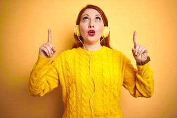 Young redhead woman listening to music using headphones over yellow isolated background amazed and surprised looking up and pointing with fingers and raised arms.