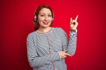 Sticker - Young redhead woman listening to music using headphones over red isolated background very happy pointing with hand and finger to the side