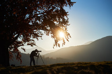 Silhouette Of Tourist Photographer Using Tripod And Professional Camera To Take Picture Of Mountain Panorama At Sunset, Standing Under Large Tree On Foggy Mountains Landscape And Blue Sky Background.