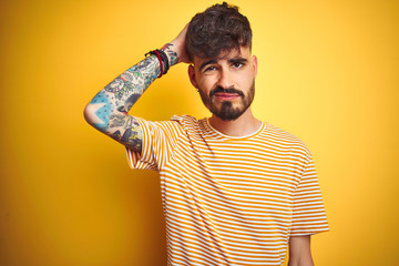 Sticker - Young man with tattoo wearing striped t-shirt standing over isolated yellow background confuse and wondering about question. Uncertain with doubt, thinking with hand on head. Pensive concept.