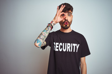 Sticker - Young safeguard man with tattoo wering security uniform over isolated white background doing ok gesture shocked with surprised face, eye looking through fingers. Unbelieving expression.