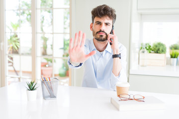 Poster - Young business man talking on smartphone with open hand doing stop sign with serious and confident expression, defense gesture