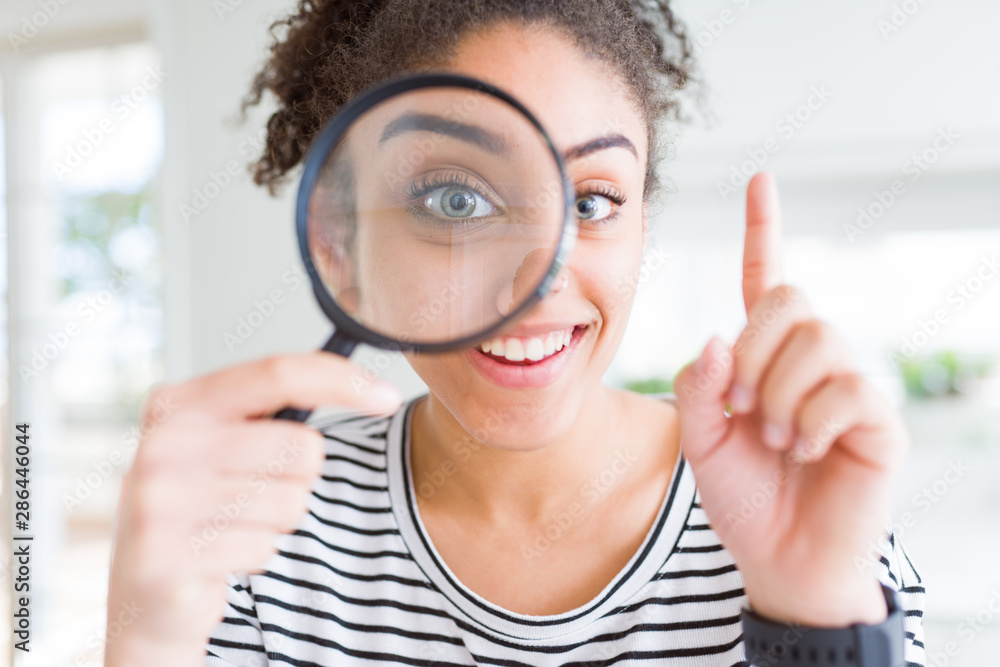 Young african american woman looking through magnifying glass surprised with an idea or question pointing finger with happy face, number one - obrazy, fototapety, plakaty 