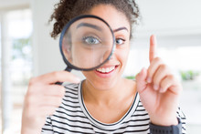 Young African American Woman Looking Through Magnifying Glass Surprised With An Idea Or Question Pointing Finger With Happy Face, Number One