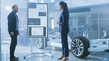 Poster - Male and Female Design Engineers are Working and Discuss Electric Car Benefits on an Interactive Whiteboard Next to an Electric Car Chassis Prototype. High Tech Laboratory Facility with Vehicle Frame.