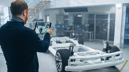 Aufkleber - Male Engineer is Working in a High Tech Development Facility Holding a Tablet Computer. He Stands Next to an Electric Car Chassis Prototype with Batteries and Wheels.