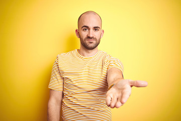 Sticker - Young bald man with beard wearing casual striped t-shirt over yellow isolated background smiling cheerful offering palm hand giving assistance and acceptance.