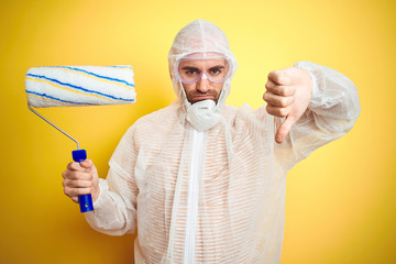 Sticker - Young man wearing painter equipment and holding painting roller over isolated yellow background with angry face, negative sign showing dislike with thumbs down, rejection concept