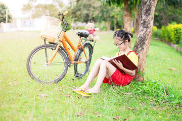 Poster - Cute asian student girl with glasses sitting on the grass reading a book in garden at summertime