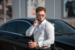 © ianachyrva - Young successful businessman in a white shirt and sun glasses drinks coffee from disposable cup and speaks on the phone standing near his luxary car.