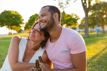 Wall Mural - Happy couple enjoying outdoor date at sunset. Man and woman sitting on grass, talking, laughing, hugging and drinking. Romance concept