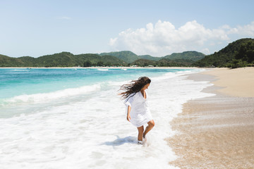 Young happy woman enjoying on tropical white sand empty beach. White tunic, long curly hair, asian brunette.