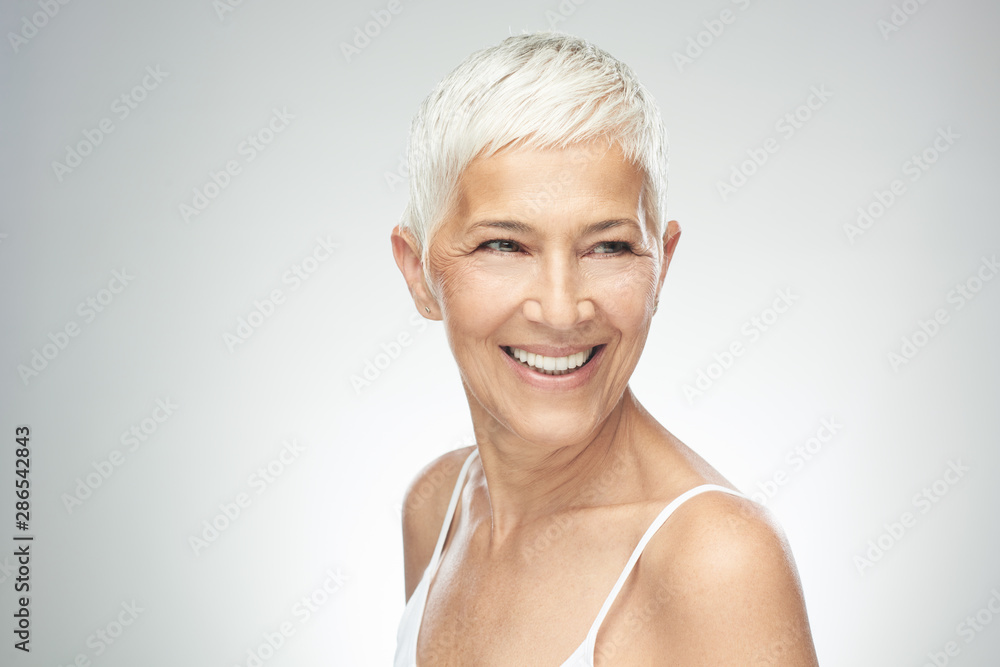 Beautiful smiling senior woman with short gray hair posing in front of gray background. Beauty photography. - obrazy, fototapety, plakaty 