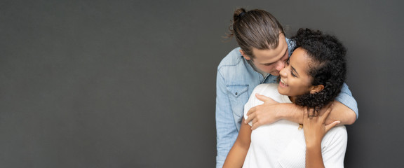 Panoramic banner image of a sweet couple hugging and kissing his wife on the cheek on black background in studio. wide crop