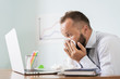 © Juliaap - Sick man with handkerchief sneezing blowing nose while working in office, businessman caught cold, seasonal flu. Pandemic influenza, disease prevention, air conditioning in office cause sickness