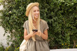 Outdoor portrait of cute young blond female in casual linen dress over green garden, keeping smartphone in hands and looking aside with broad smile