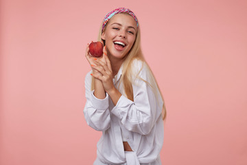 Wall Mural - Young lovely blonde woman with orange manicure in white shirt looking to camera and holding peach near cheek, being positive and happy, isolated over pink background