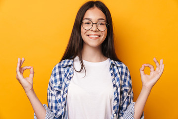 Wall Mural - Pleased optimistic young cute teenage girl in glasses meditate isolated over yellow wall background.