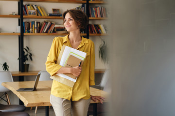 Aufkleber - Young attractive woman in yellow shirt leaning on desk with notepad and papers in hand while dreamily looking aside in modern office