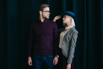 Wall Mural - stylish young man in glasses and blonde woman in hat looking at each other near curtain