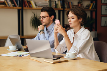 Aufkleber - Young woman sitting at the table with laptop and cup of coffee thoughtfully using cellphone. Business colleagues working in office