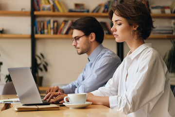 Aufkleber - Young serious woman sitting at the table tiredly working on laptop with colleague near in office
