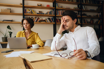 Aufkleber - Young exhausted man sitting at the table with notepad and eyeglasses while tiredly holding hand near face at work in office with colleague on background
