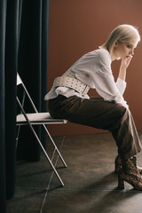 Wall Mural - side view of stylish blonde woman in white blouse and boots with snakeskin print sitting on chair near curtain on brown