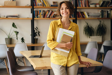 Young cheerful woman in yellow shirt leaning on desk with notepad and papers in hand while joyfully looking in camera in modern office