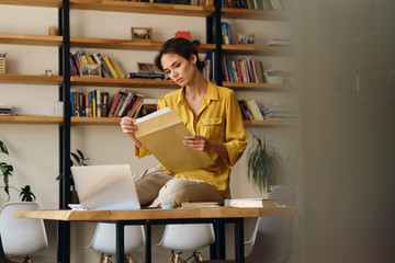 Aufkleber - Young beautiful woman in yellow shirt sitting on desk with papers and laptop while thoughtfully working in modern office