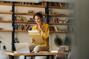 Wall Mural - Young attractive woman in yellow shirt sitting on desk with laptop while dreamily working with papers in modern office