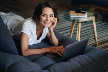 Wall Mural - Young beautiful smiling woman lying on sofa with laptop joyfully looking in camera in office